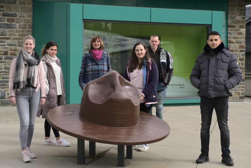 Schülerinnen und Schüler stehen vor einer großen Bronzeplastik in Form eines Hutes und hören gespannt zu